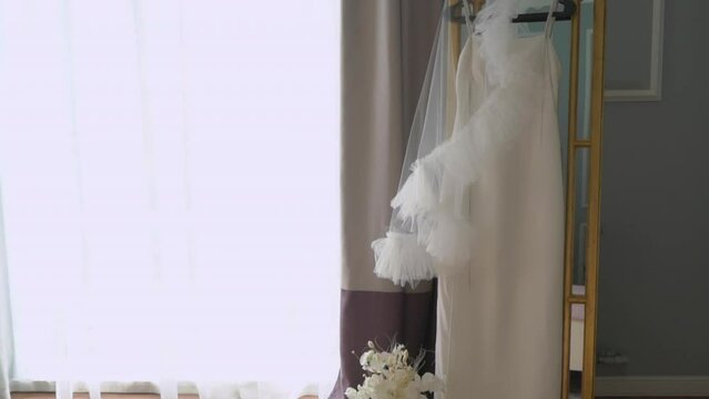 A beautiful white wedding dress and veil hangs on a hanger on the mirror in the bedroom. Morning of the bride, wedding day. Women's fashionable clothes for the holiday.