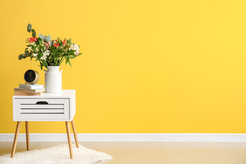 Vase with beautiful alstroemeria flowers, alarm clock and books on end table near yellow wall