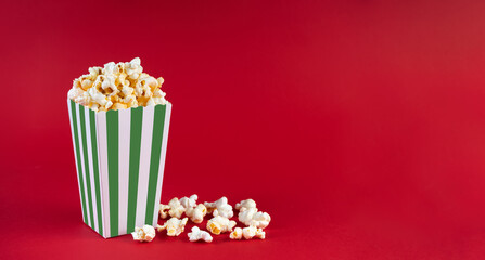 Green white striped carton bucket with tasty cheese popcorn, isolated on red background. Box with scattering of popcorn grains. Fast food, movies, cinema and entertainment concept.