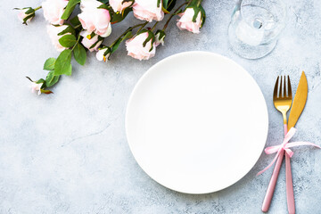 Table setting with white plate, modern cutlery and pink rose flowers. Flat lay at white with copy space.