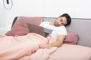 Lazy young handsome man sleeping on bed while working on laptop at home