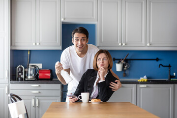 Tired business woman sits at the table and her husband householder takes care about her in the kitchen