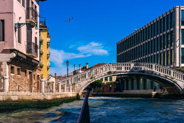 A stone bridge over the canal on Venice Street next to tall and vintage buildings on a sunny summer day, authentic Italian architecture, panoramic view, a gull in the blue sky over the city