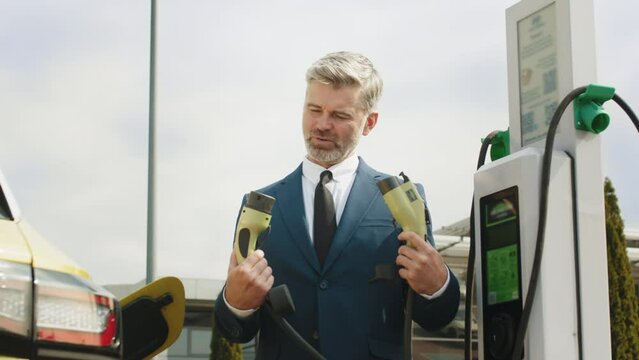 Handsome mature grey haired thoughtful man at a loss holding two power cable for environmentally friendly zero emission electric car. Businessman charging electric car at outdoor charging station.