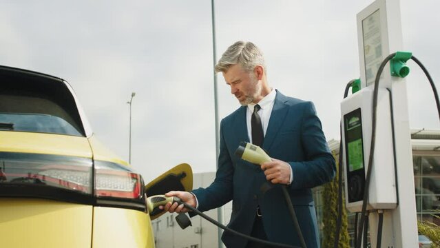 Problem with charging an electric car, different connection cables. Handsome mature thoughtful man at a loss holding two power cable for electric car. Businessman charging electric car