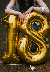 A girl on her birthday holds in her hands airy foil golden balloons with the number 18 in the park....