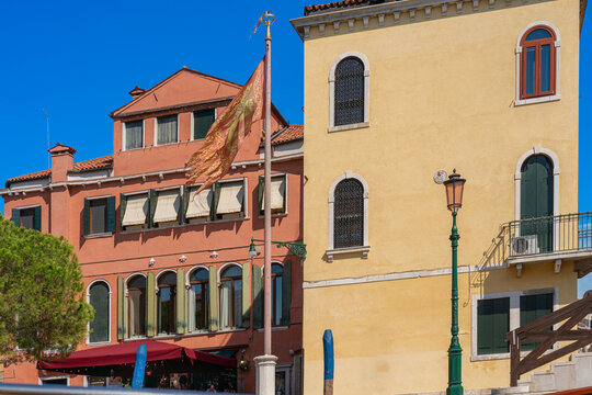 Venetian flag on a high pole against the background of the walls of old ancient houses with high windows and wooden shutters, blue sky, summer sunny day, authentic Italian architecture, real world