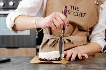 A young, smiling pastry chef in a beige apron prepares a small cake with a offset spatula in a modern kitchen. Close up.