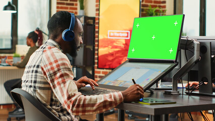 Young content creator using greenscreen background on computer, retouching photos with editing software. Male employee working with isolated chroma key display and blank copyspace.