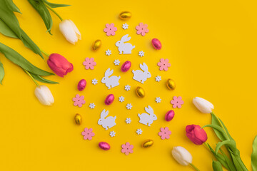 Easter flat lay with white bunny cutouts, chocolate eggs, pink flowers, and tulips on a bright yellow background. A festive spring composition for holiday designs and decorations.