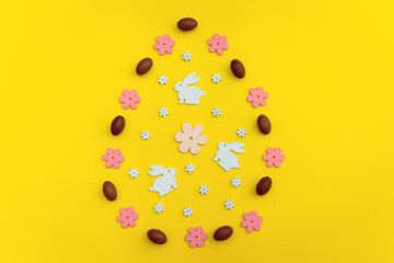 Easter flat lay with white wooden bunny cutouts and pink flowers, chocolate eggs,  arranged in an egg shape on a yellow background. A festive spring concept for holiday designs and decorations.