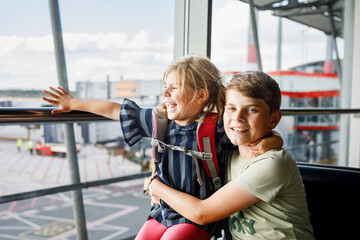 Little girl and school boy at the airport waiting for boarding at big window. Two kids stands at window against the backdrop of airplanes. Happy children, siblings leaving for family summer vacation