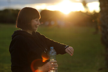 Mature adult woman with bottle of water looking at smart watch to check exercise progress after morning routine on nature. Senior healthy lifestyle, vitality, healthcare concept. World health day