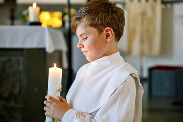 Little kid boy receiving his first holy communion. Happy child holding Christening candle. Tradition in catholic curch. Kid in a white traditional gown in a church near altar.