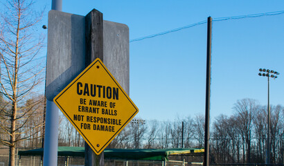 Caution be aware of errant balls not responsible for damage street sign in a parking lot by a baseball field