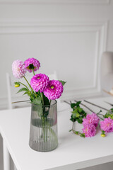 Vertical image of pink dahlias standing in a transparent vase on a white table. The concept of greetings to women's day, a place for your design