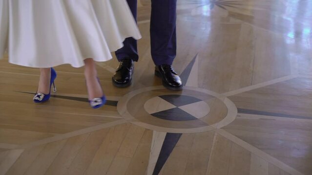 The bride in a long wedding dress is spinning, tearing the hem of her dress. Slowmotion indoors. Blue heeled shoes, groom in suit.