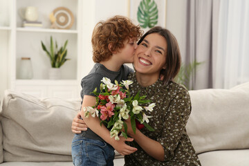 Little son kissing and congratulating his mom with Mother`s day at home. Woman holding bouquet of...