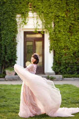 Fototapeta premium Beautiful woman in flying airy pink dress on background of door overgrown with wild grapes. Pretty young caucasian girl plays with her dress. Girl dance in flowing dress