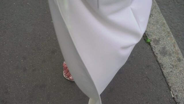 A young woman in a white dress and high-heeled sandals shoes is standing on the street. The hem of the dress flutters in the wind. Bride in wedding day outdoors.
