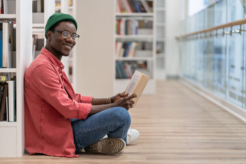 Black student man library visitor sitting on floor near bookcase with book in hands, looking at camera and smiling, African American guy literature lover in public library interior. Reading hobby