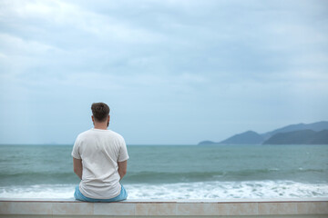 Young calm sad serious man is sitting on embankment near sea, ocean on beach, thinking, meditate on...