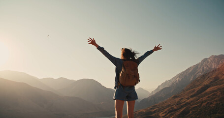 Young girl standing on top of mountain and victoriously raising hands up, looking far away....