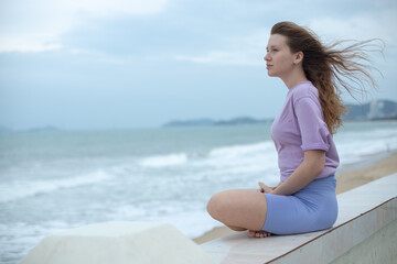Young calm sad serious woman is sitting on embankment near sea, ocean on beach, girl thinking,...