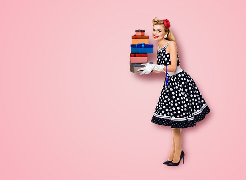 Big sales discounts, rebates offers ad concept - full body image of happy smiling beautiful woman in pin up black dress with polka dot, white gloves, hold gift boxes, isolated rose pink background