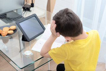 School boy in yellow t-shirt sitting at the table with digital tablet studying at home. Distance learning online education concept