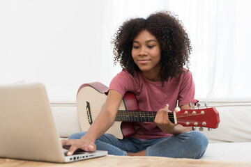 Happy young woman playing guitar on sofa and using laptop computer in living room at home