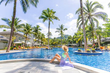 Young unrecognizable woman relaxes by the edge of a beautiful swimming pool, enjoying a moment of peace and tranquility at the luxurious spa resort. Asia tropical vacation. - obrazy, fototapety, plakaty