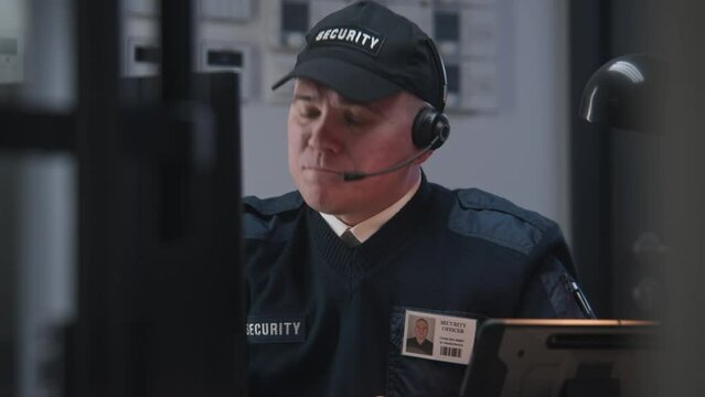 Serious security officer in headphones sits on workplace and looks view from security camera displayed on monitor. Man talks with coworker by walkie talkie. Tracking systems. CCTV technology. Zoom in.