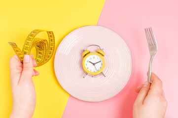 Female hands holding measuring tape and fork near empty plate on yellow and pink background top...