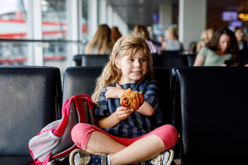 Little girl at the airport waiting for boarding at the big window. Cute kid eating bun for a snack....