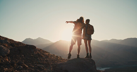 Young couple with backpack standing on the top - happy people enjoying summer holidays at the mountains- Millennial woman and man holding hands showing mountains together.. Freedom concept - obrazy, fototapety, plakaty