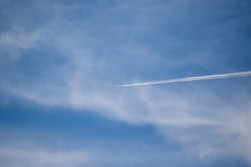 Vapor trail from a jet flying high overhead dissipates into the thin white clouds in a blue sky