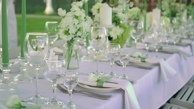 Served tables for a holiday in a restaurant. Glasses, plates, cutlery, napkins. Wedding decor, celebration dinner or party, reception indoors. No food. White and green candles outdoors in summer.