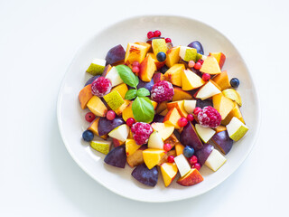 Delicious fruit salad with fresh plum, peach, nectarine, pear and frozen raspberries, decorated with green basil on white background. Top view. Healthy eating. Copy space. Vegan cuisine. Dietary menu.