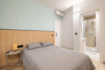 Fototapeta na wymiar Double bed with gray linen, small bedside table and wooden slats on the wall overlooking compact shower room. Concept of small multifunctional apartment. Copyspace