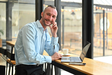 Obraz premium Smiling millennial man using laptop, working remotely in bright coffee shop interior. Business, freelance concept