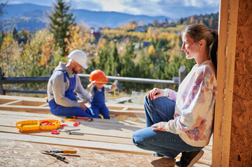 Father, mother and son building wooden frame house. Toddler boy helping his daddy, while woman looking for them on construction site. Guys wearing helmet and blue overalls. Carpentry, family concept.