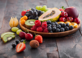 Fresh raw organic summer berries and exotic fruits in round wooden plate on dark wooden kitchen background. Papaya, grapes, nectarine, orange, raspberry, kiwi, strawberry, lychees, cherry.