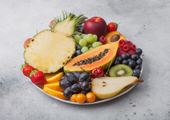 Fresh raw organic summer berries and exotic fruits in white plate on light background. Pineapple, papaya, grapes, nectarine, orange, apricot, kiwi, pear, lychees, cherry and physalis.