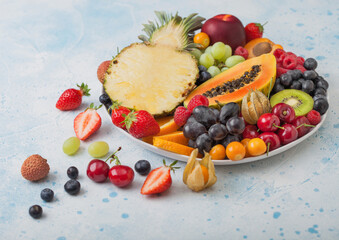 Fresh raw organic summer berries and exotic fruits in white plate on blue kitchen background. Pineapple, papaya, grapes, nectarine, orange, apricot, kiwi, pear, lychees, cherry and physalis.