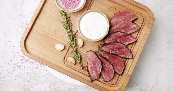  Sliced Beef Meat. Rare medium cooked beef with garlic and rosemary, white sauce and salt in a Woden plate. The salt falls on the meat. Restaurant Food. Rotating Plate on white background
