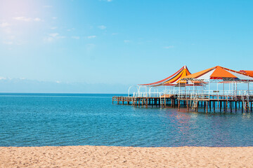 Wooden pier with orange-red awning over water of sea, lake, ocean with beach without people. Travel, vacation, summer background. Copy space