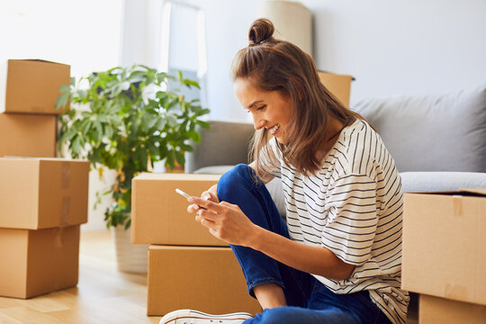 Beautiful young woman texting while moving to new apartment