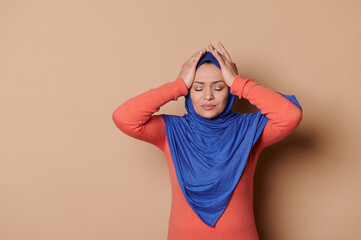 Unhealthy Middle-Eastern Muslim woman in blue hijab, rubbing temples to cure headache problem, suffering from tension, migaine stress, grimacing in pain, high blood pressure, isolated beige background - obrazy, fototapety, plakaty