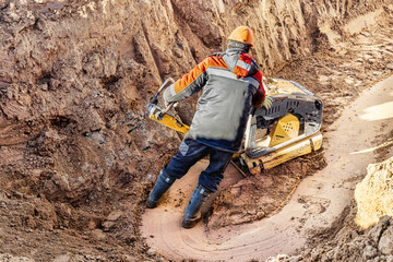 A worker compacts soil or sand with a vibrating plate in a trench at a construction site close-up....